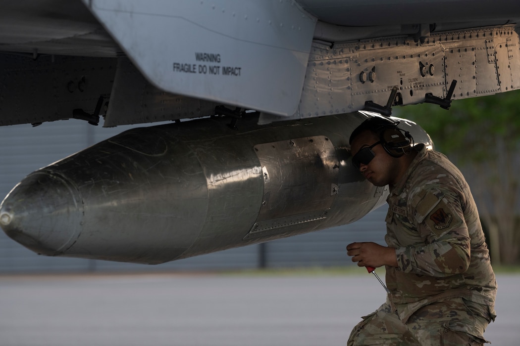 A person crouching underneath an A-10C, looking at a bolt on a travel pod.