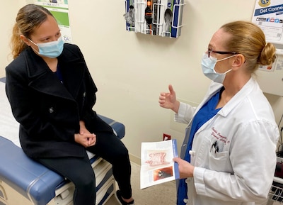 Patient wearing mask sits on exam table and speaks with doctor, wearing mask and lab coat, who is holding a pamphlet about colon health.