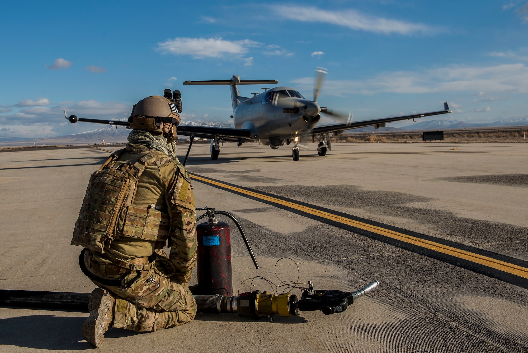 A person in military uniform kneels on a concrete runway in front of a single engine military plane.