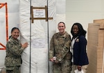 Three women stand in front of an object covered in white shipping paper.