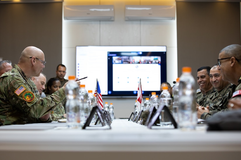 Service members in uniforms with various camouflage patterns sit across from each other at a table that has miniature flags and water bottles on top of it. One of the service members is pointing at another one across the table who is smiling.