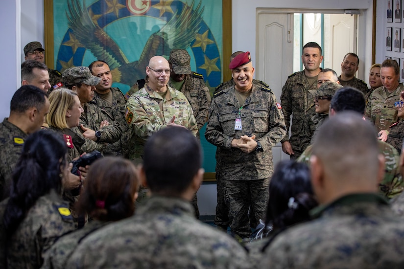 A group of about 20 service members in uniforms with various camouflage patterns are inside a room, standing in a circle and talking to each other.