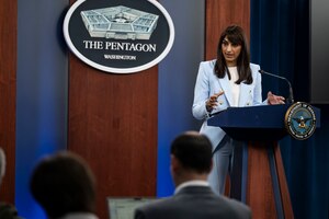 A woman stands at a podium talking to a couple of people sitting down with their backs to the camera. There is a sign behind the woman that has a five-sided building on it, and that reads “The Pentagon, Washington.”