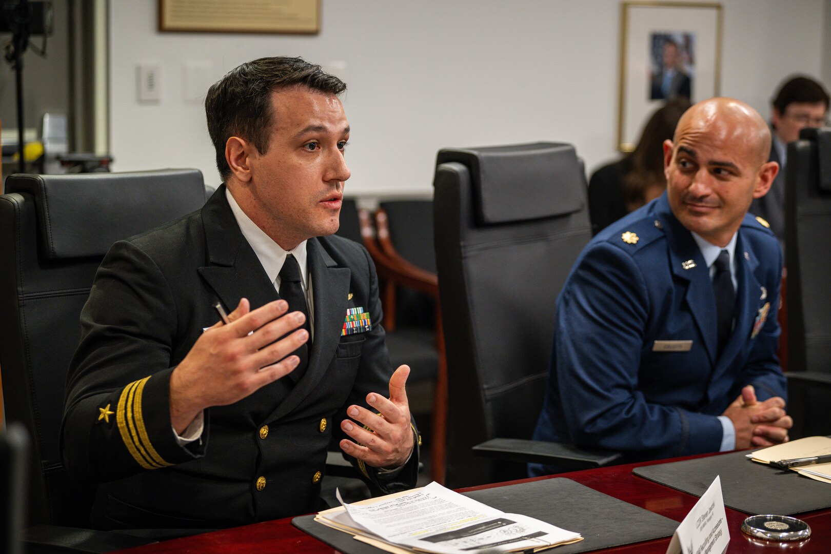 A service member listens while seated next to another who gestures while speaking.