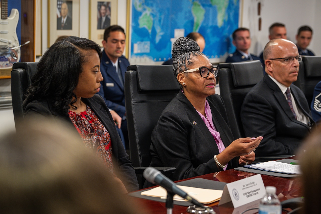 A speaker gestures while surrounded by other seated individuals.