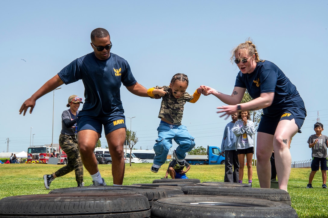 A child jumps above a tire obstacle in a field outdoors while service members on either side hold the child's hands.