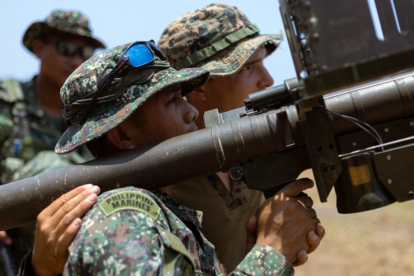 A Philippine Marine shoulder aims a weapon system while standing next to a U.S. Marine outdoors, with a third service member watching.
