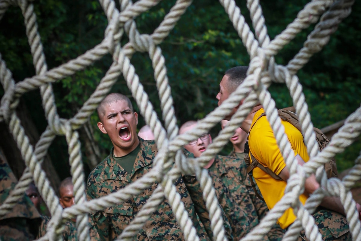 A group of Marines stand behind a thick rope obstacle. One Marine is yelling.