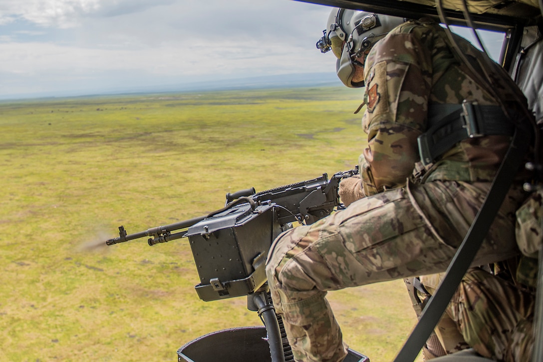 A uniformed service member wearing eye and head protection operates a machine gun through the open section of a helicopter during daytime training over a field.