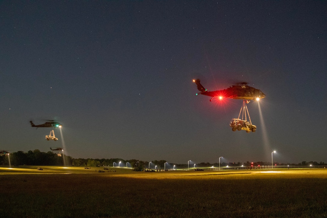 Two military helicopters carry large military vehicles by sling load at night.