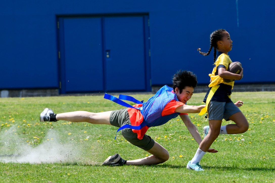 A student falls to the grass while reaching for the flag of another child who is running with a football.