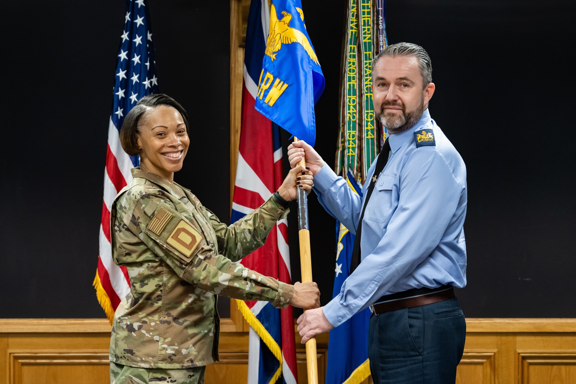 U.S. Air Force Chief Master Sgt. Tiffany Griego, left, 100th Air Refueling Wing command chief, presents Royal Air Force Warrant Officer Paul ‘Jerry’ McGuire, RAF Honington station warrant officer, with the 100th ARW guidon during an honorary commander induction ceremony at RAF Mildenhall, England, April 24, 2024.