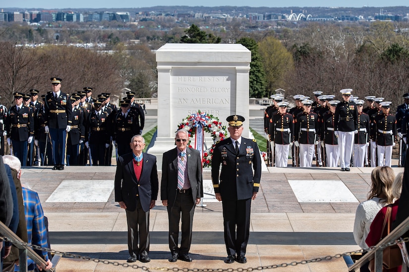 Two people in business suits stand with a uniformed service member at attention in front of a monument. Several others in uniform stand at attention behind them.