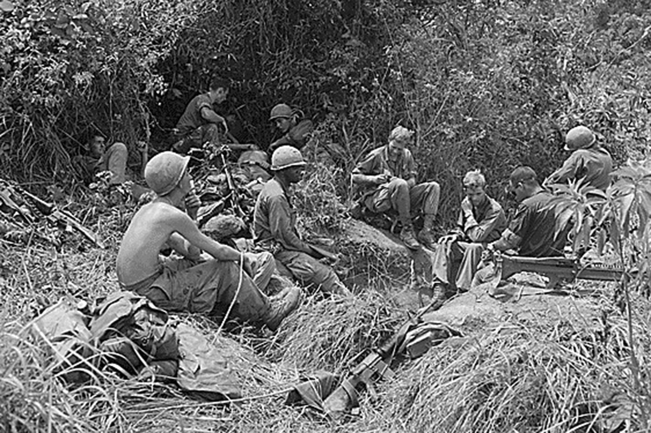 Several soldiers relax in tall grass near dense foliage.