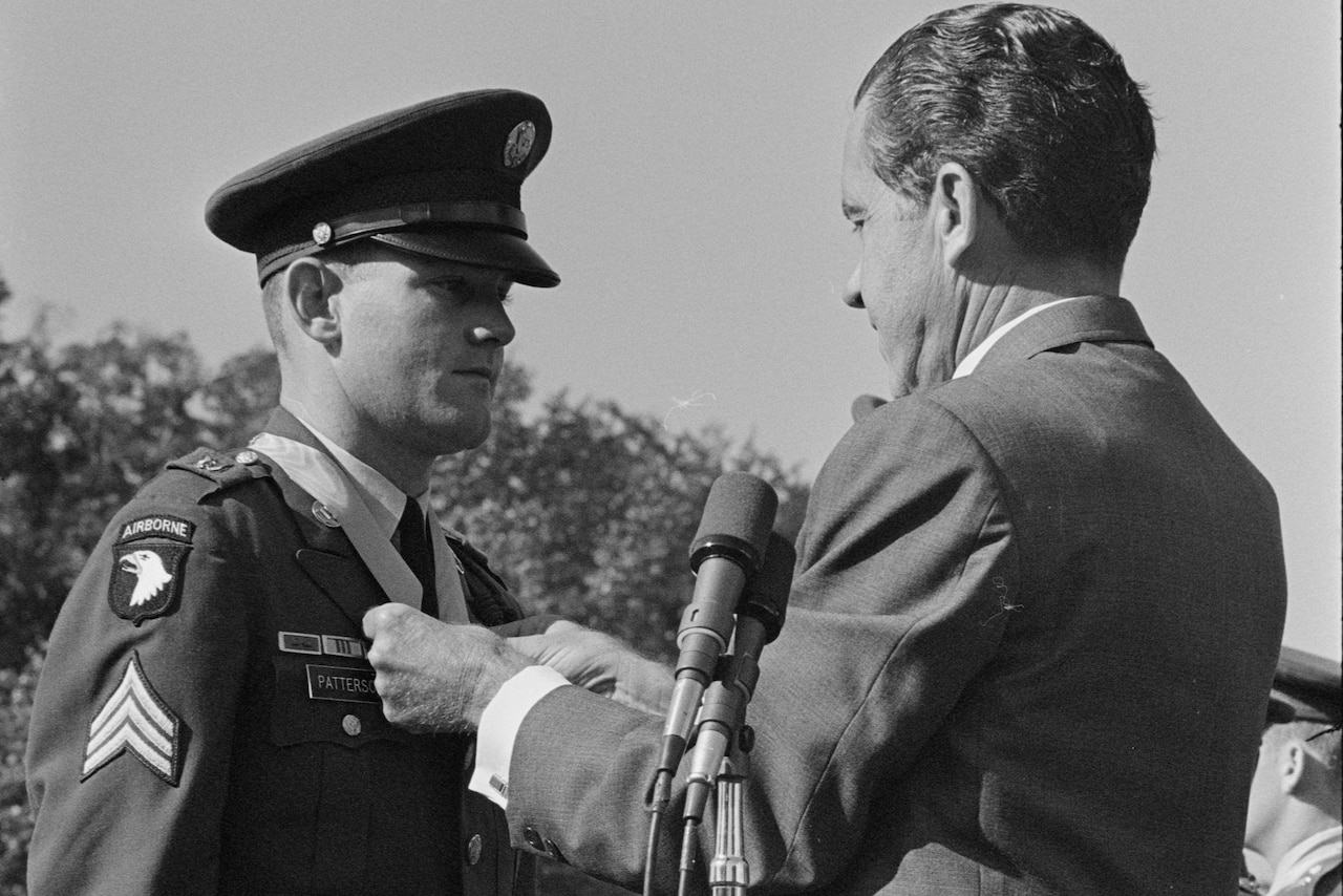 A person in a business suit places a ribboned medal around a uniformed service member's neck.
