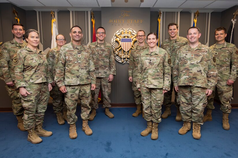 A group of uniformed service members pose for a photo in front of flags.