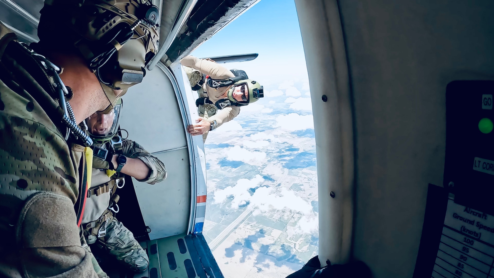 A man hangs onto the side of the plane in midair, preparing to jump with two others inside the plane.