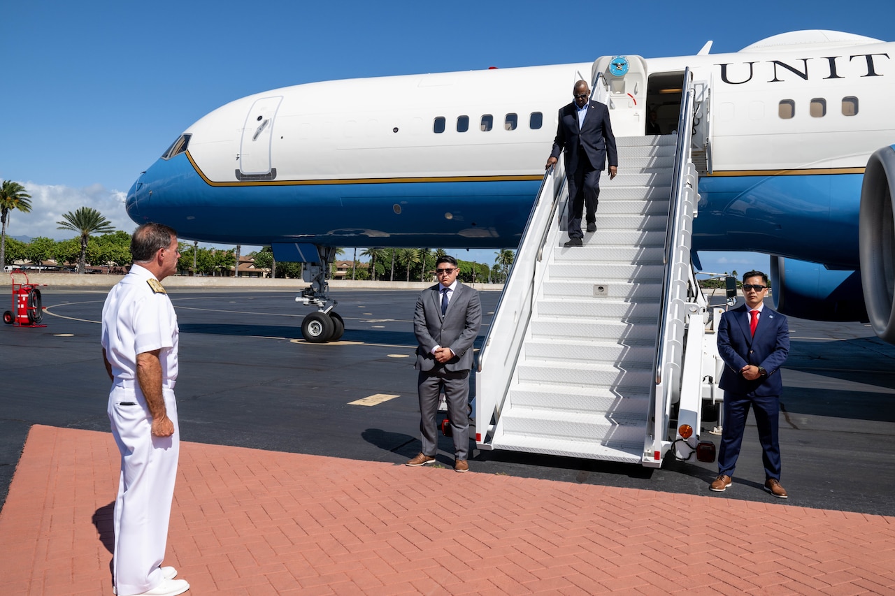 A service member in uniform stands at attention as a civilian wearing a suit disembarks an airplane.