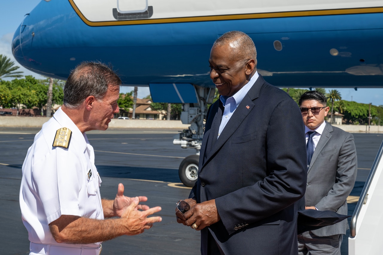 A service member in uniform and a civilian man wearing a suit engage in conversation.