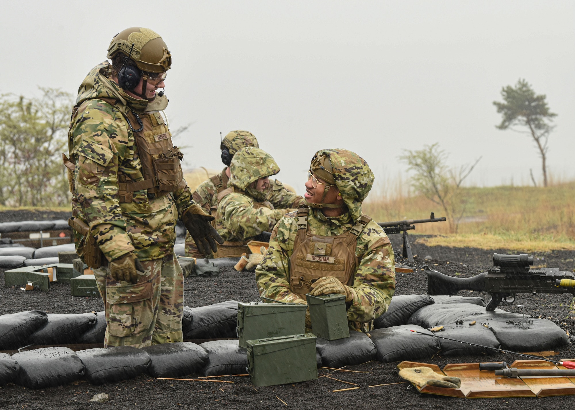 Two U.S. servicemembers speak while other members prepare to fire a machine gun in the background.