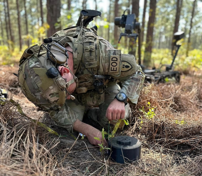 A kneeling soldier manipulates a puck-shaped device on the ground in a forested area.