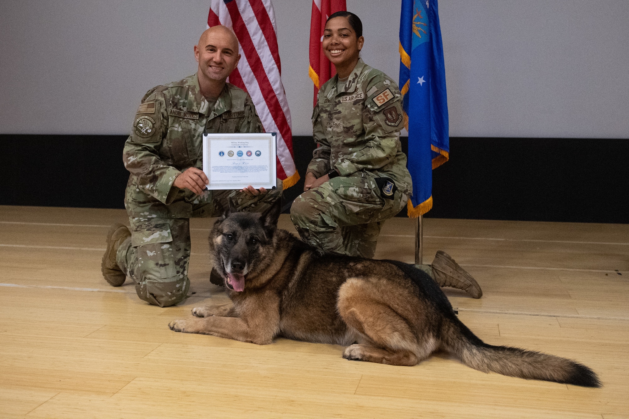U.S. Air Force Capt. Alexander Andrejkovics and Staff Sgt. Ivana Glover, kneel on the ground while holding MWD Brix's retirement certificate. Brix is laying on the ground.