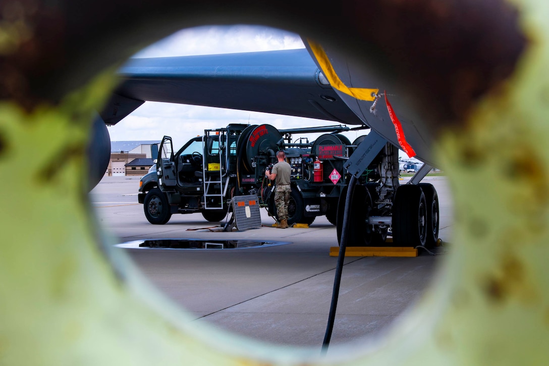 An airman is seen through a round hole refueling a large aircraft on a tarmac.
