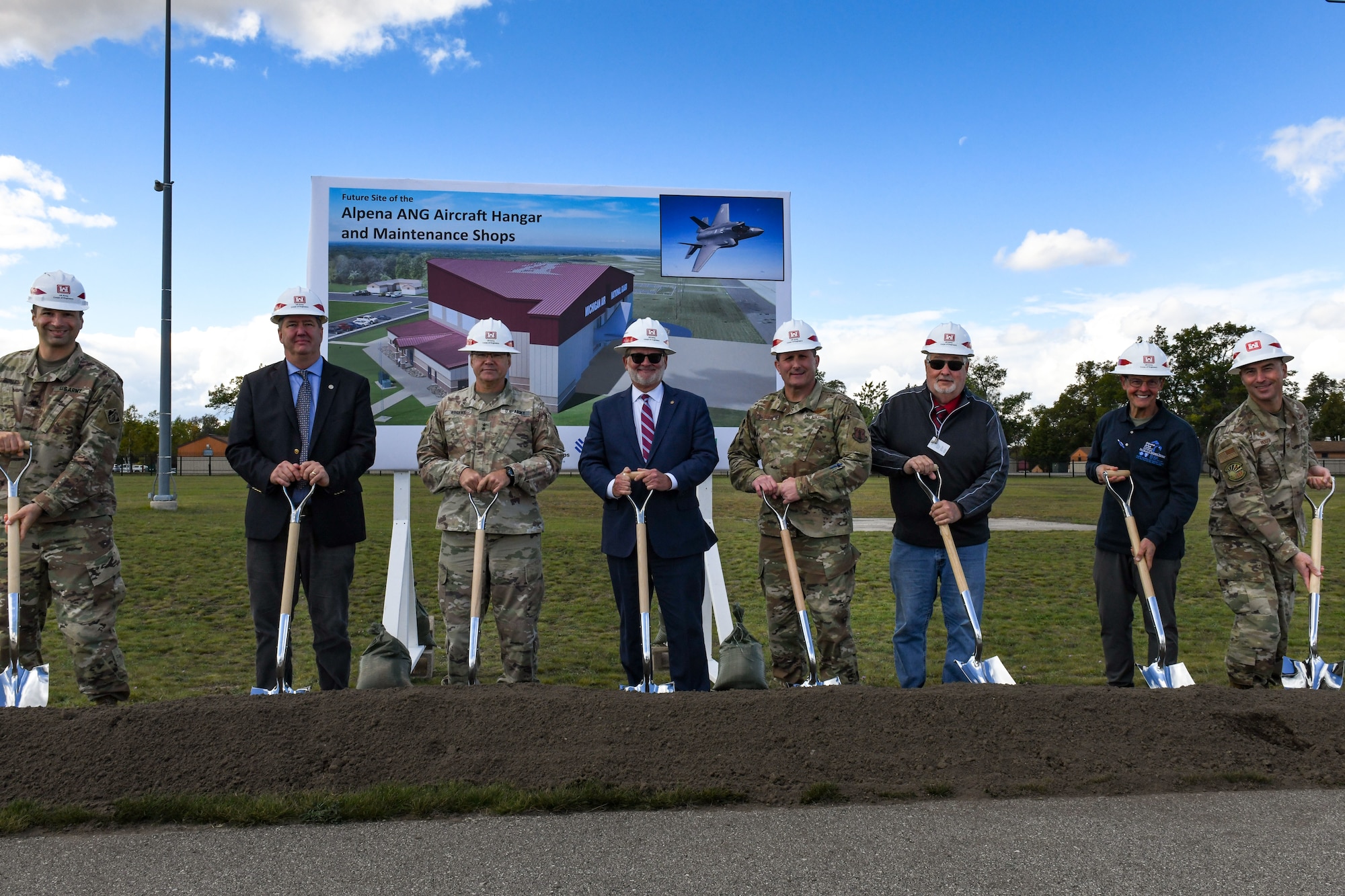 Line of people in military uniforms and business suits, all wearing hard hats and holding shovels in dirt.