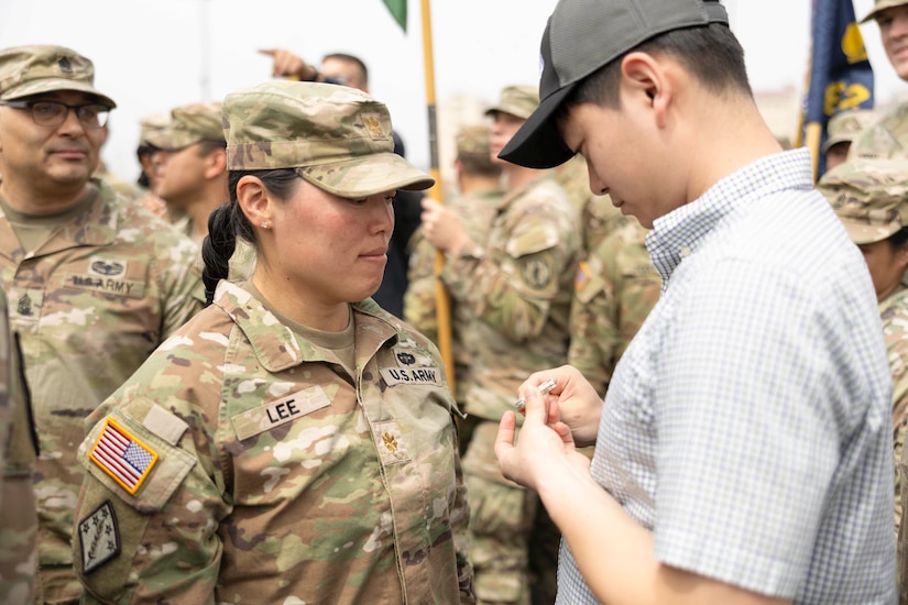 A military spouse holds a pin before putting it onto an emotional soldier as fellow soldiers stand in the background.