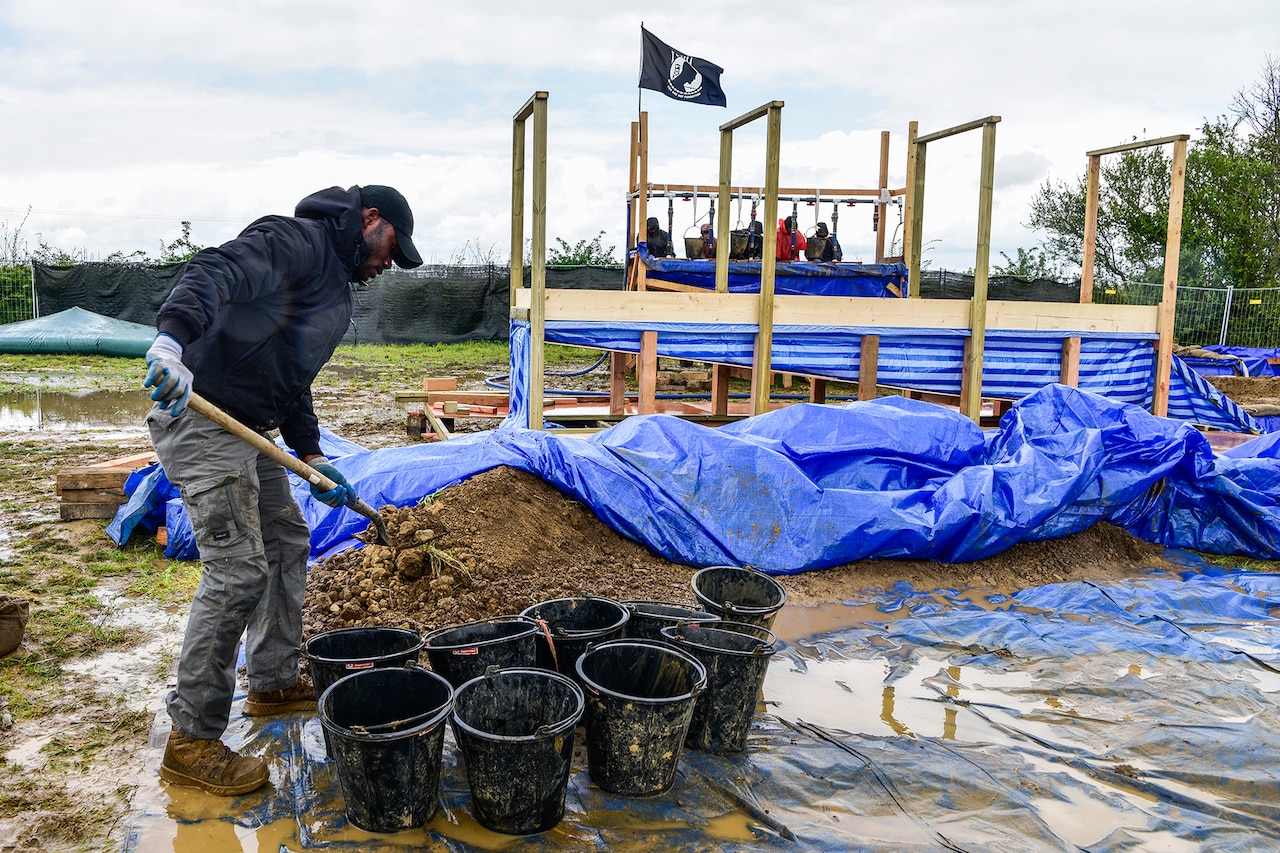 A person uses a shovel to fill several black buckets with dirt. Blue tarp can be seen covering some of the soil.