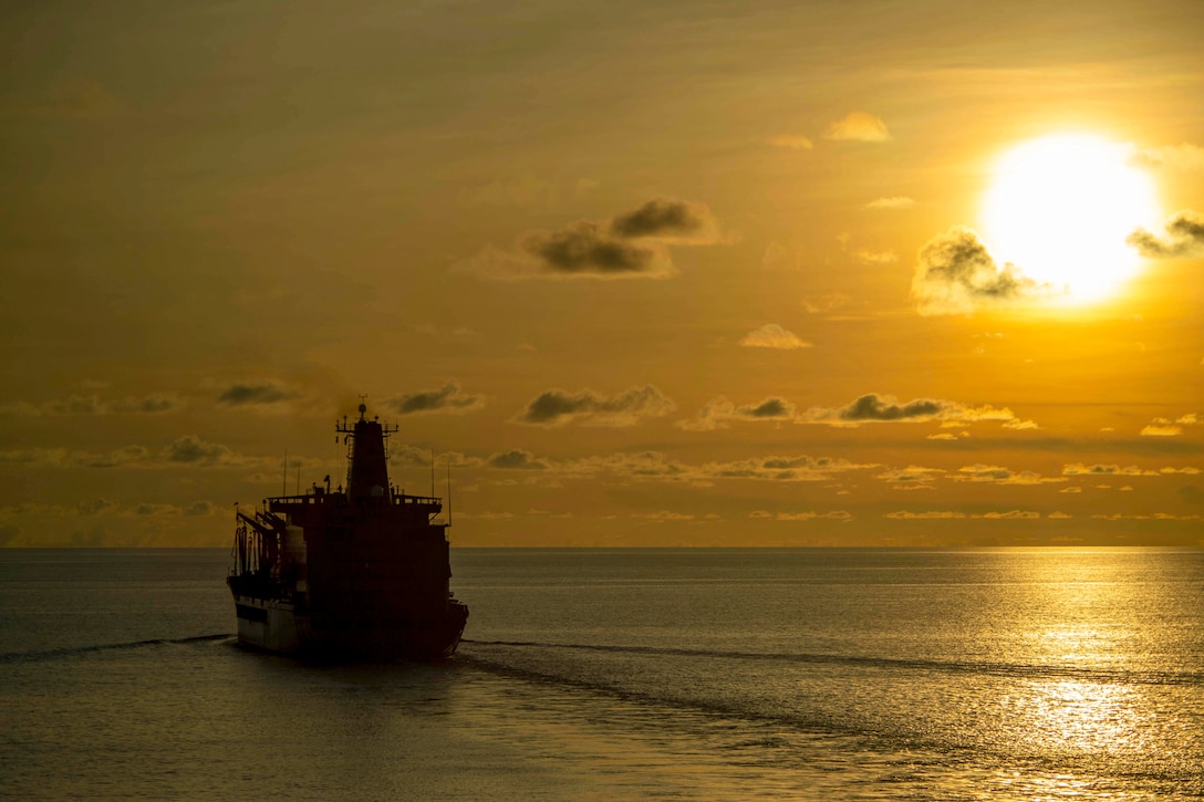 A ship in silhouette sails in a body of water under a sunlit sky.