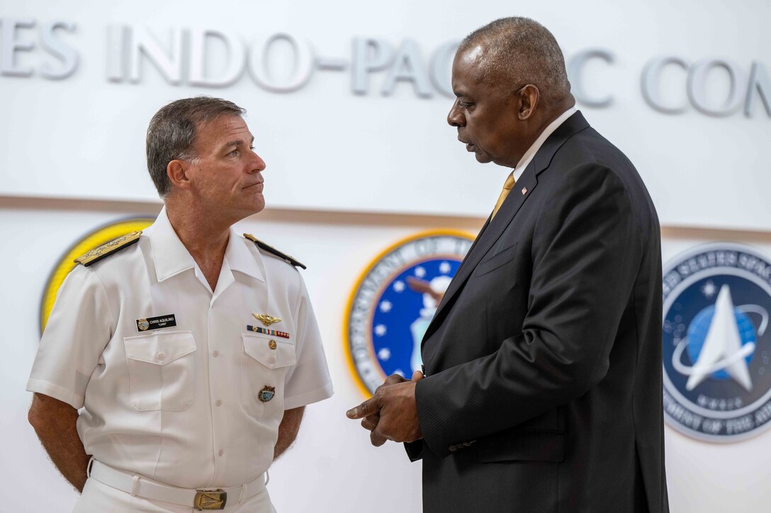 A service member in uniform and a civilian in wearing a suit stand face to face in conversation.