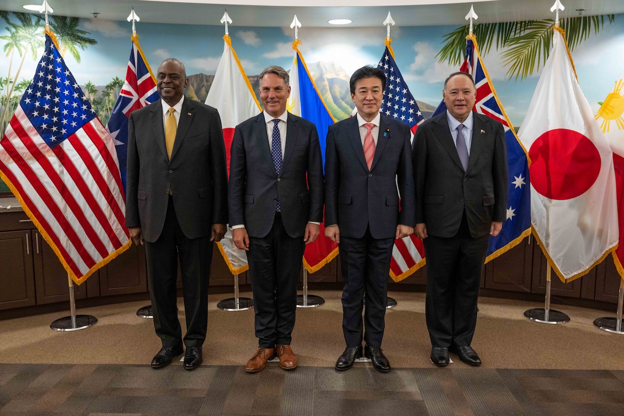 Civilians wearing suits stand side by side in front of U.S., Australian, Japanese and Filipino flags.