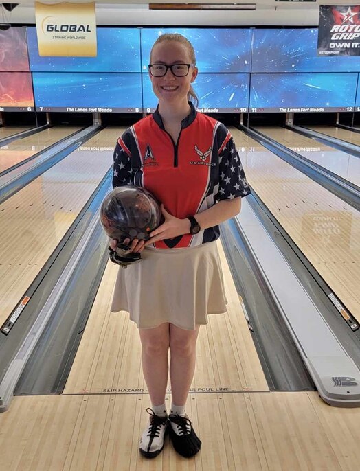  Woman in bowling uniform posing for a photo holding a bowling ball in front of bowling lanes