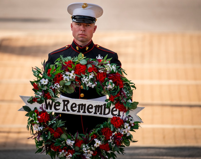A Marine in dress uniform holds up a floral wreath with a sign reading "We Remember" in the middle.