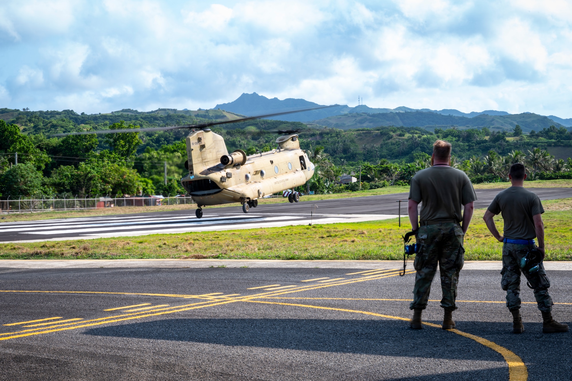 Two individuals stand watching an army aircraft takeoff.