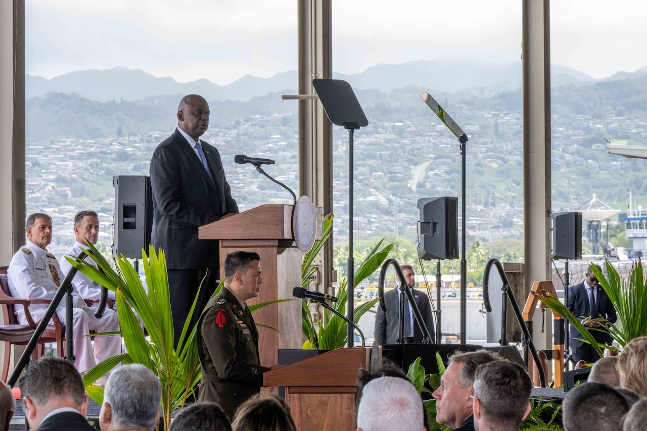 A civilian speaks from a stage in front of an audience.