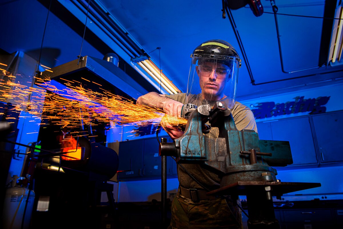 An Air Force cadet wearing protective head and eye gear forges a knife, emitting bright orange sparks.