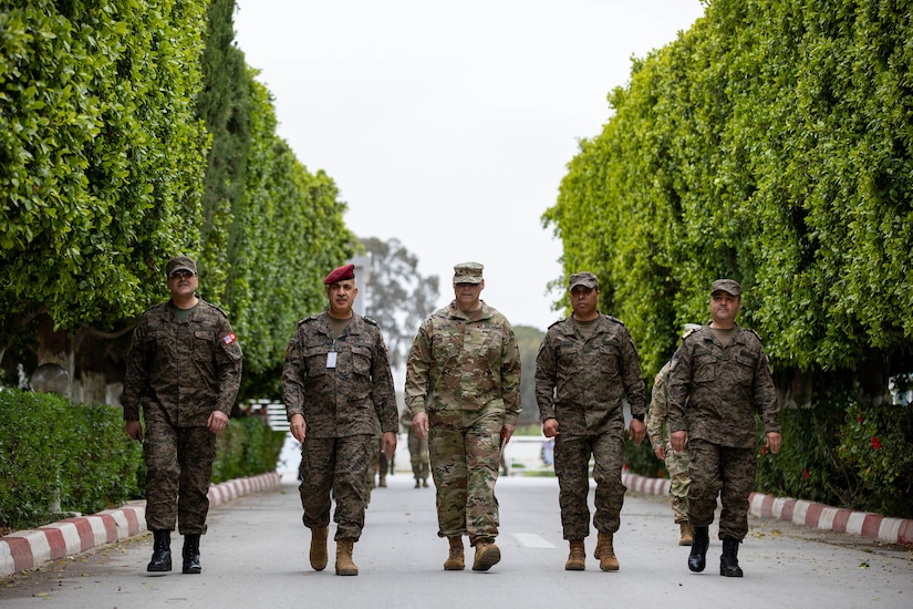 Five people in military uniforms walk side by side, towards a camera.