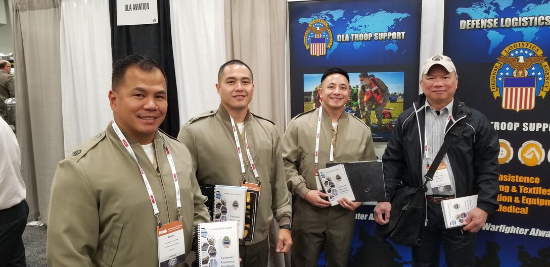 Four men - three in khaki jackets - stand in front of poster displays holding a ringed book.