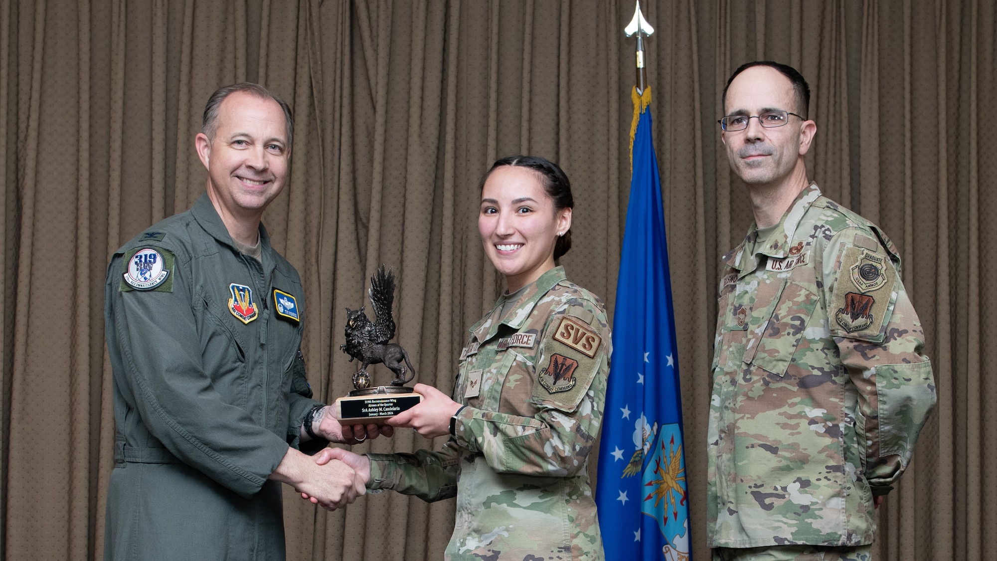 Two men and one woman  in military uniforms stand on stage exchanging an award.