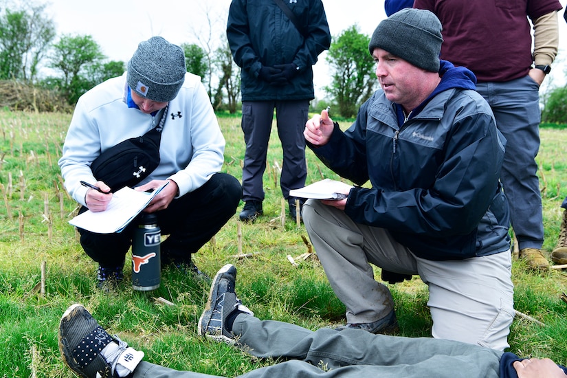 Two people write on notepads while a person lays on the ground and others stand behind them.