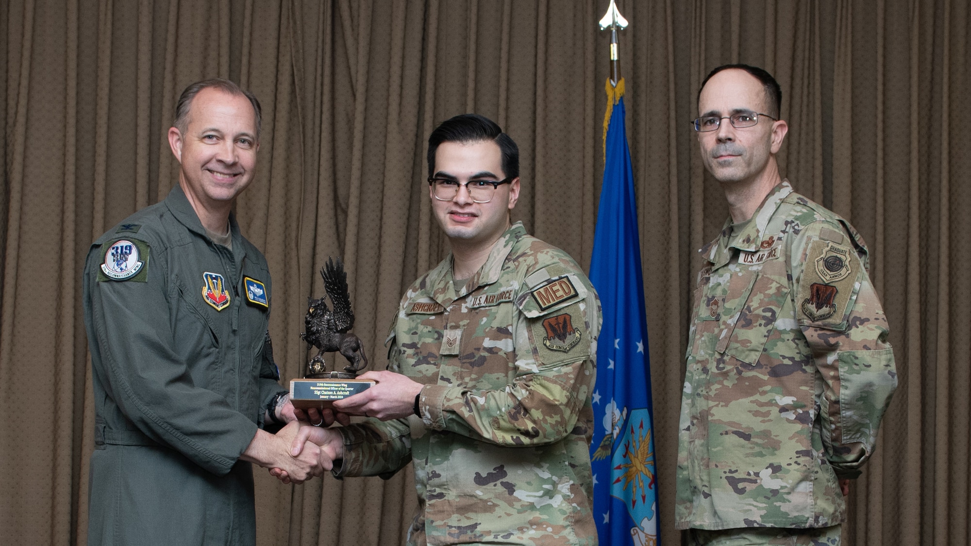Three men in military uniforms stand on stage exchanging an award.