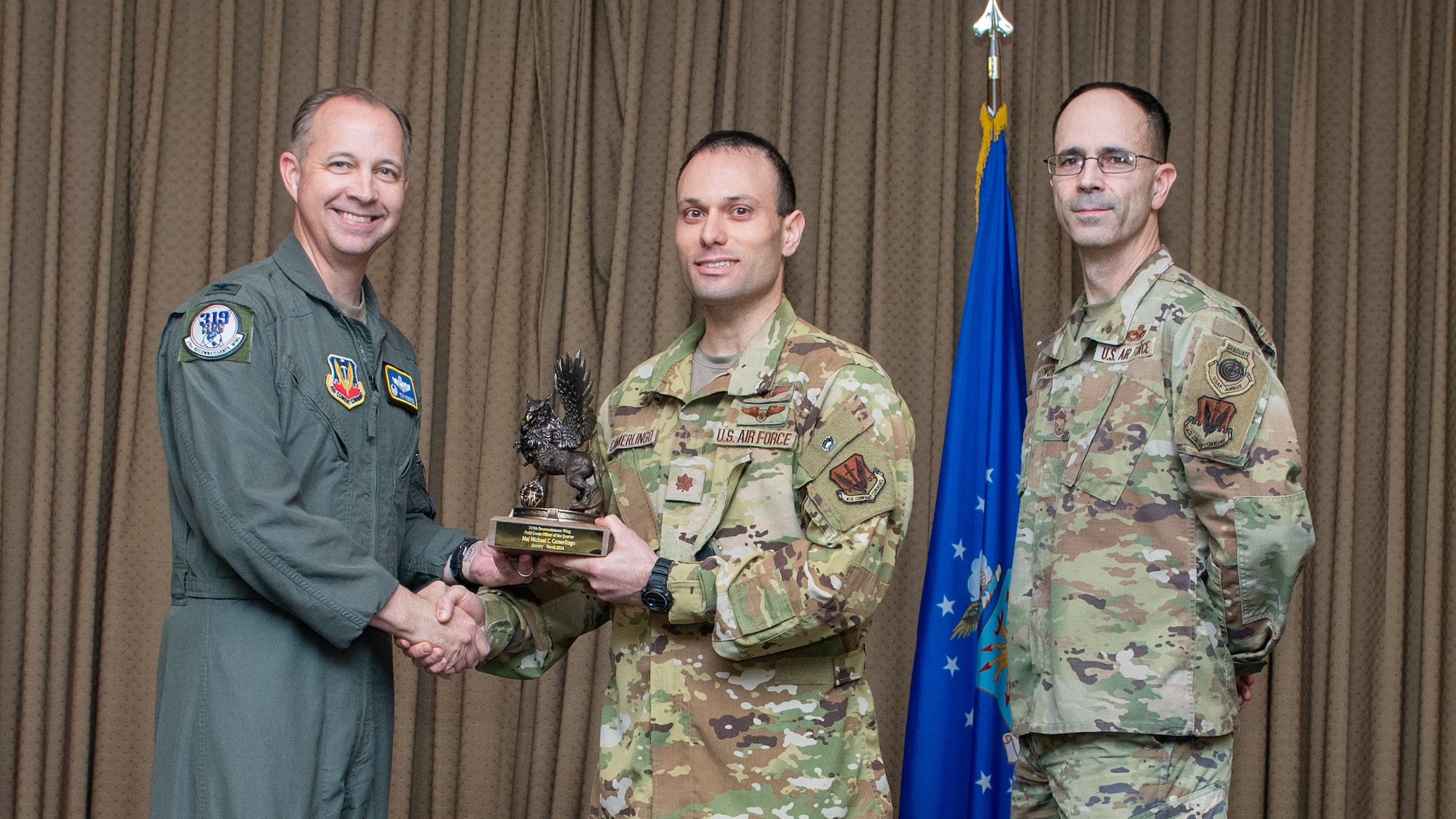 Three men in military uniforms stand on stage exchanging an award.