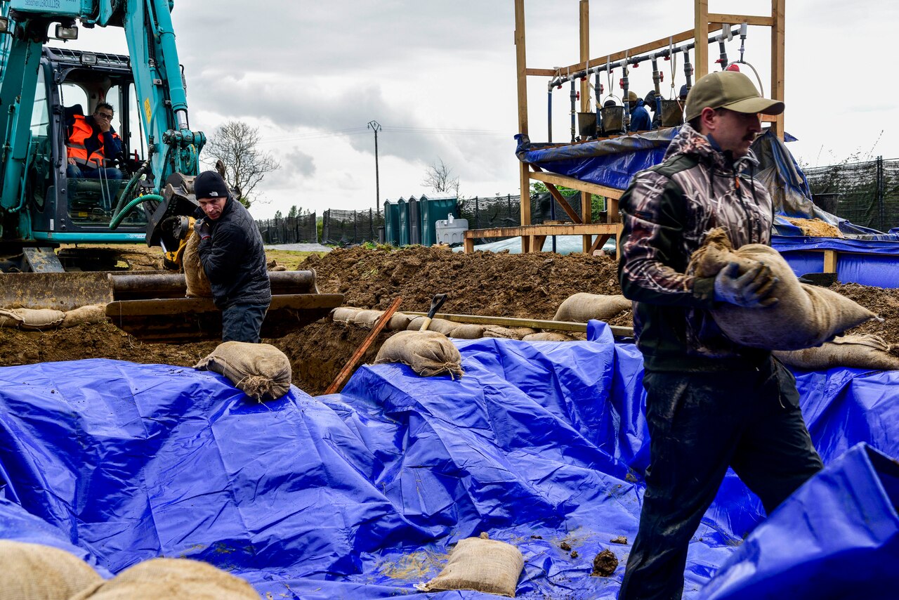Two people fill sandbags while another digs dirt with an excavator machine.