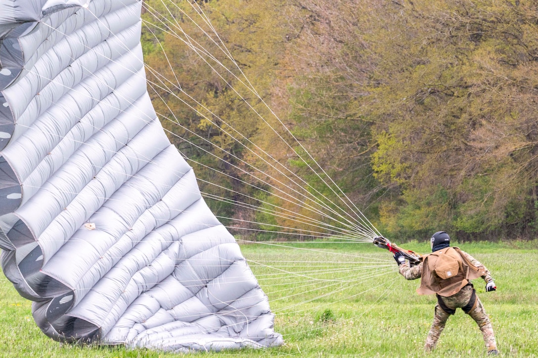 A soldier collapses their parachute while standing in a field with trees in the background.