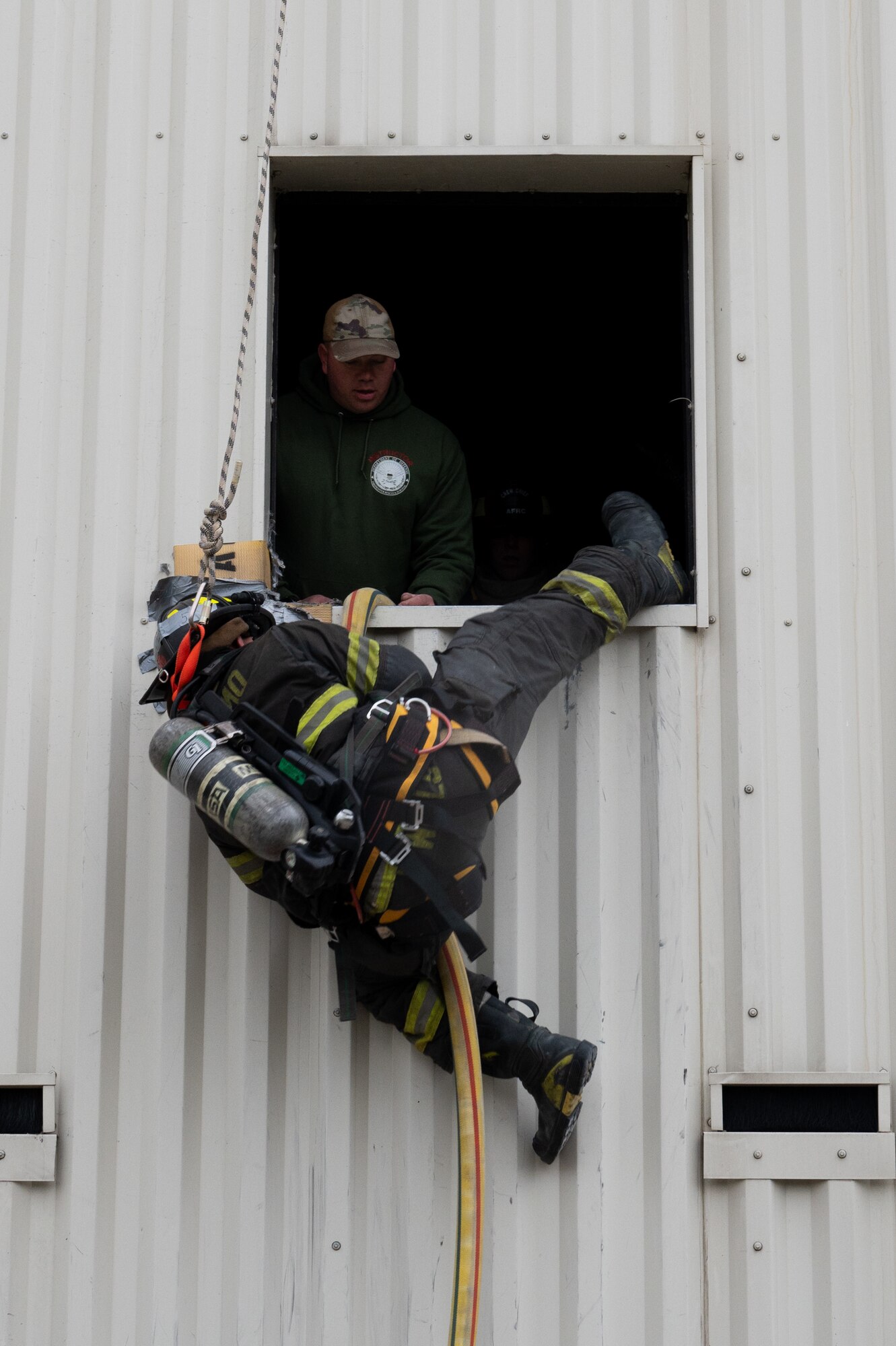 A firefighter exits a window via fire hose as another firefighter observes from the window.