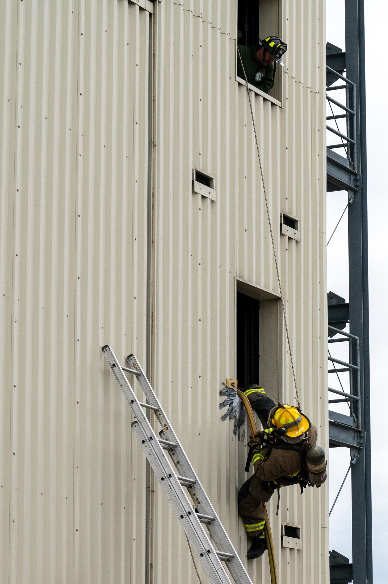 A firefighter bailing out of a window while another leans out of an above window to observe.