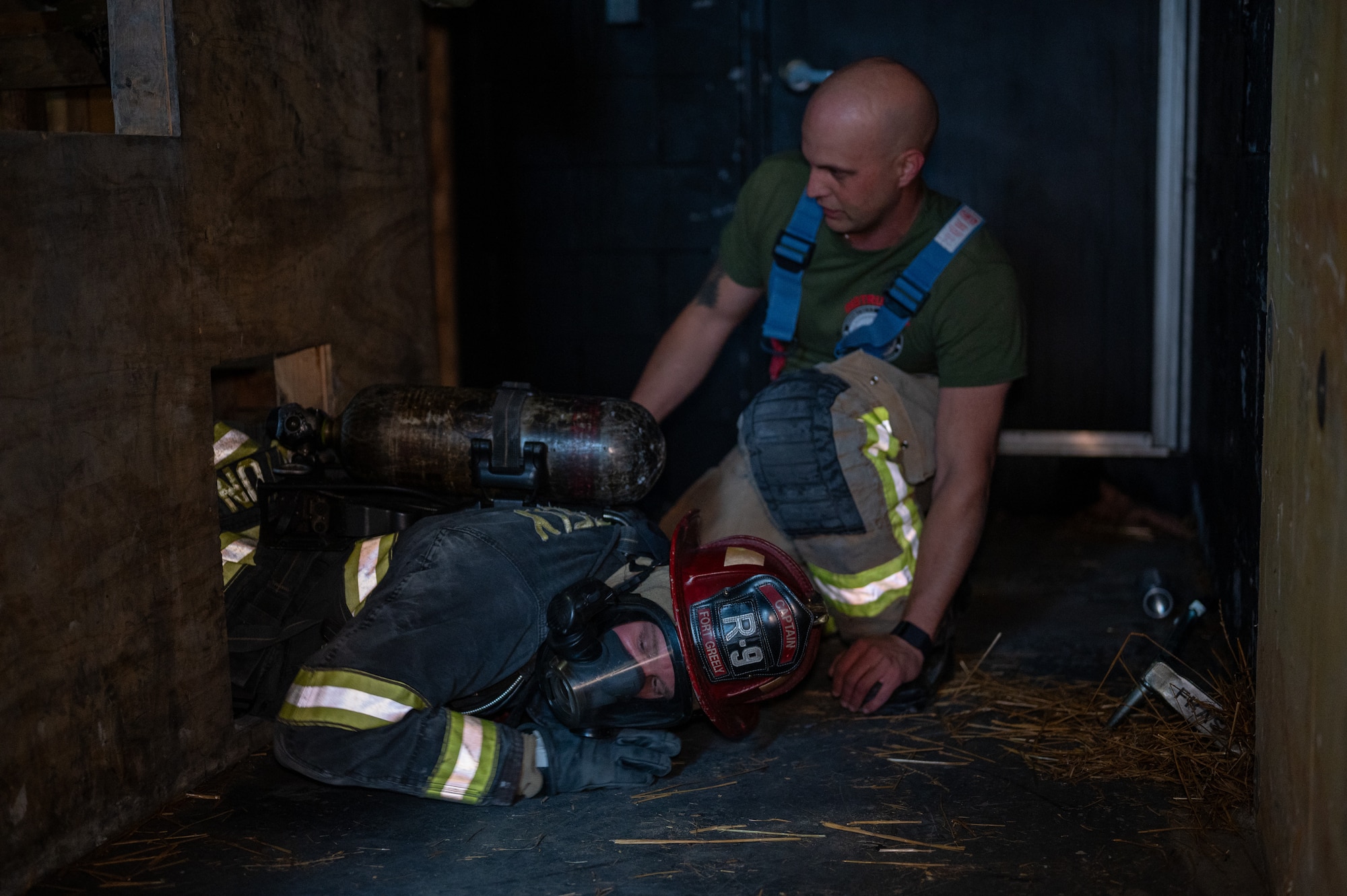 A firefighter crawls out of a confined space. In the background, another man observes his technique.