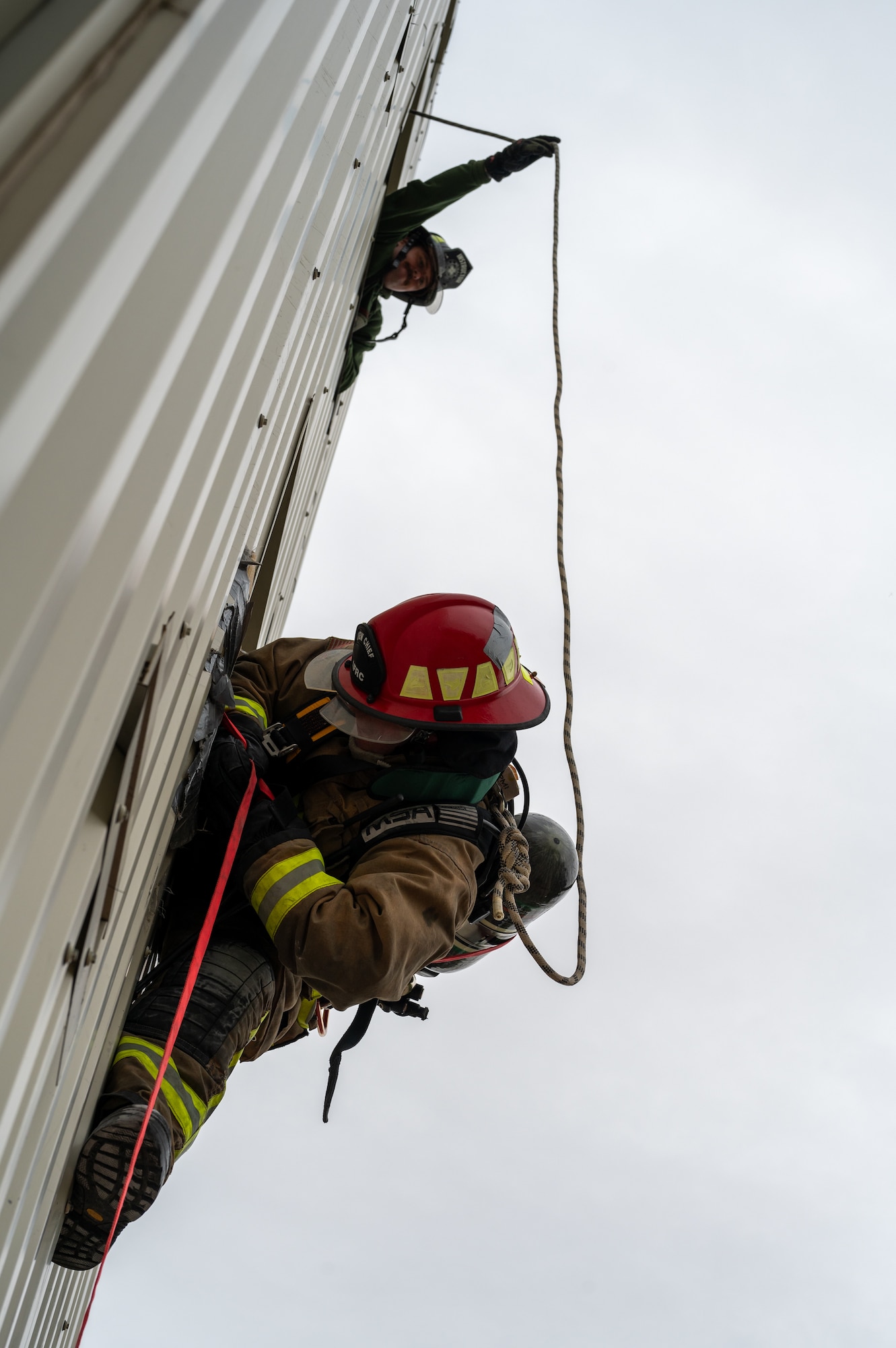 A firefighter leans out of a window while another above holds the belay line away from the exterior of the building.